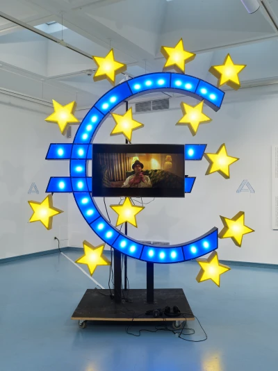 Alex Wissel und Jan Bonny:
Eurozeichen (Rheingold), 2018, 
Fiberboard, Acrylglas, 
Aluminium, Bildschirm, Video
Courtesy: Kunsthaus NRW Kornelimünster
